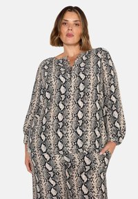 Leichte Bluse mit Schlangenprint in Creme und Schwarz, rundem Ausschnitt, Rüschenverzierungen und langen Ärmeln. Mit seitlichen Taschen.