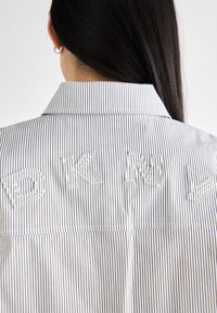 Pruhovaná košile na knoflíky s bílým vyšitým nápisem "DKNY" na zádech. Tkanina je lehká s hladkou texturou a klasickým límcem.