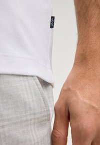 Wit katoenen T-shirt met een klein marineblauw label met de tekst "JOOP!" aan de zijkant, gecombineerd met lichtgrijze, gestructureerde broek. Close-up van hand en kleding.