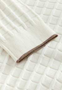 Manche de pull en tricot blanc avec bordure brune et côtes, reposant sur un fond en tissu à mailles blanches texturées.
