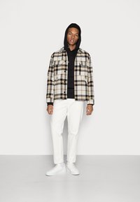 Karierter Blazer in Beige und Schwarz über einem schwarzen Hoodie, kombiniert mit weißen Hosen und weißen Sneakern. Glattes Material, Knopfverschluss und zwei Taschen.