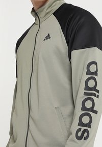 Homme portant une veste zippée Adidas gris clair et noire avec le logo Adidas sur la poitrine et un grand texte sur la manche.