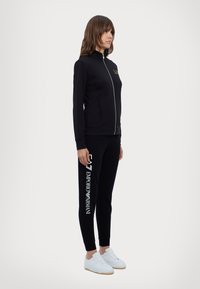 Musta vetoketjullinen takki ja housut, joissa on valkoisella teksti "Emporio Armani" housuissa. Täydentää asun valkoiset tennarit. Sileä kankaan tekstuuri.