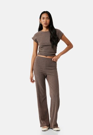 Vrouw met lang zwart haar die een bijpassende bruine top met korte mouwen en wijde broek draagt, staand met één hand op haar heup en witte schoenen.