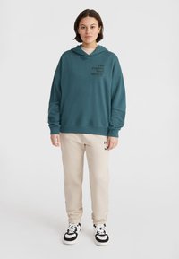 Teal capuchon sweatshirt met zwarte tekst, gecombineerd met beige sweatpants met merklogo. Model staat op zwarte en witte sneakers.