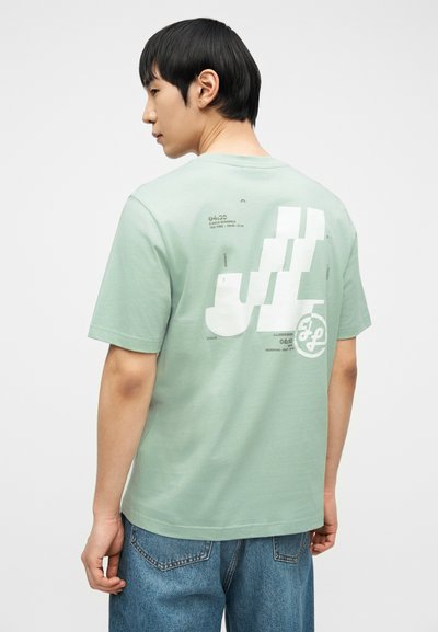 Άνδρας που φοράει ανοιχτοπράσινο μπλουζάκι με λευκό γραφικό κείμενο στην πλάτη και μπλε τζιν, στέκεται μπροστά σε απλό ανοιχτό φόντο.