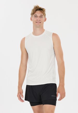 Jeune homme debout, portant un débardeur blanc de sport et un short de course noir à couches, souriant vers la caméra.