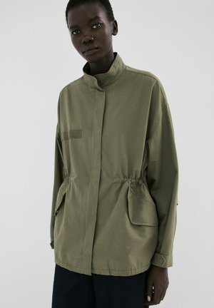 Persona de piel oscura que lleva una chaqueta verde oliva de gran tamaño con un cuello alto y grandes bolsillos delanteros, de pie frente a un fondo blanco liso.