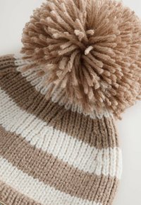 Stickad mössa med växlande bruna och vita ränder, prydd med en stor fluffig pom-pom på toppen. Materialet verkar mjukt och texturerat.