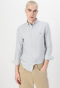 Lichtblauw en wit gestreept overhemd met knopen en een kraag, lange mouwen opgerold bij de manchetten, met een klein logo op de borst.