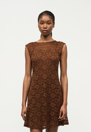 Joven con el cabello trenzado, vestida con un vestido marrón de encaje sin mangas, de pie frente a un fondo claro y liso, mirando hacia adelante.