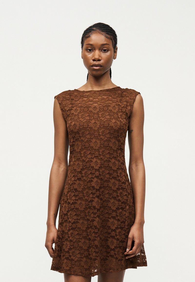 Junge Frau mit geflochtenem Haar, die ein ärmelloses braunes Spitzenkleid trägt, steht vor einem schlichten hellen Hintergrund und schaut nach vorne.