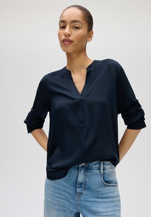 Donna con i capelli legati indossa una camicetta blu scuro con le maniche arrotolate e jeans blu chiaro su uno sfondo semplice grigio chiaro.
