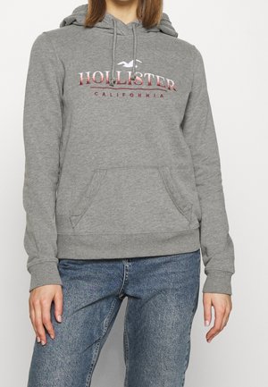 Person iført en grå Hollister California hoodie med frontlomme og blå jeans, stående foran en ensfarvet baggrund.