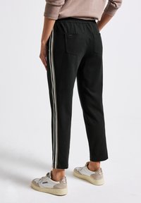 Person som bär svarta byxor med vita sidostråkar och vita sneakers, stående med en hand vilande på höften, sett bakifrån.