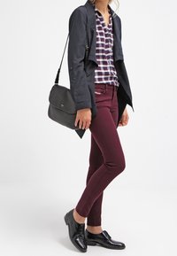 Kvinna klädd i en marinblå jacka, rutigt skjorta, vinröda byxor, svarta läderskor och bär på en svart axelväska.