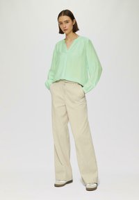Hellgrüne Bluse mit langen Ärmeln und V-Ausschnitt; beigefarbene, weite Hose mit glatter Textur und Gesäßtaschen; lässige Sneakers.