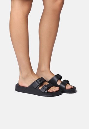 Black slide sandals made of textured material, featuring two adjustable buckles, open-toe design, and a flat sole.