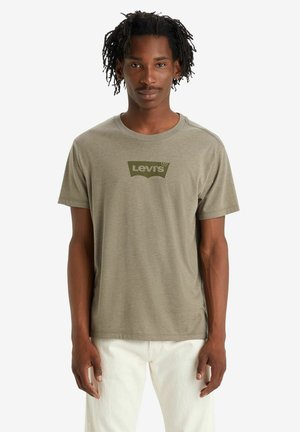 T-shirt olive à manches courtes en coton, arborant un logo Levi's vert foncé contrastant au centre, associé à un pantalon blanc.