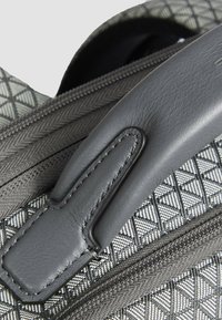 Dettaglio di una borsa grigia con motivo, caratterizzata da un manico in pelle liscia, tessuto strutturato e una cerniera con design intrecciato.