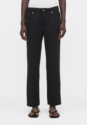 Személy fekete egyenes szárú farmert, fehér bordázott felsőt és fekete flip-flop papucsot visel, egyszerű fehér háttér előtt áll.