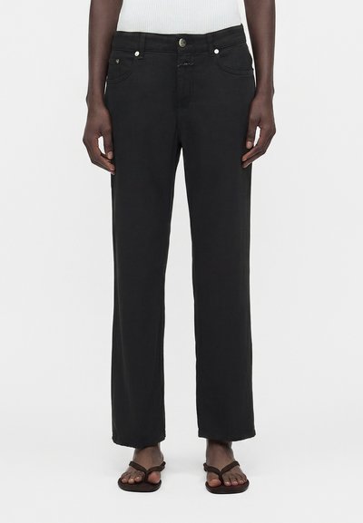 Personne portant un jean noir droit, un haut blanc côtelé et des tongs noires, debout devant un fond blanc uni.