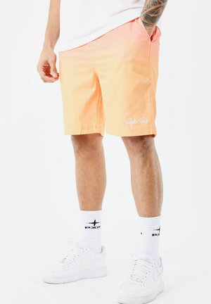 Lichtoranje zwemshorts met wit logo, elastische tailleband en zijzakken. Gecombineerd met witte sneakers en witte sokken met logo.