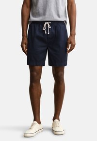 Homme portant un short bleu marine à cordon, un t-shirt gris clair et des baskets blanches, debout devant un fond uni.