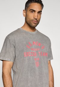 Šedé bavlněné tričko s nápisem "U.S. MEN'S NATIONAL SOCCER TEAM" červeným potiskem, krátké rukávy, kulatý výstřih a mírně texturovaný povrch.
