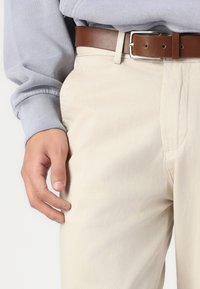 Pantalon beige clair avec une finition texturée. Assorti d'une ceinture en cuir brun, la main reposant près de la poche latérale. Un sweat-shirt gris est visible.
