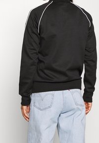 Person som bär en svart långärmad topp med vit kantning och ljusblå denimjeans, sedd bakifrån mot en vit bakgrund.