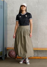 Crna košulja s uzorkom i bijelim detaljima, suknja do struka maslinaste boje, sivi tenisice i bijela kapa, držeći košarkašku loptu u mreži.