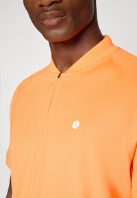 Kortermet oransje prestasjonskjorte med glidelås på kragen, teksturert stoff og en liten hvit logo på brystet.