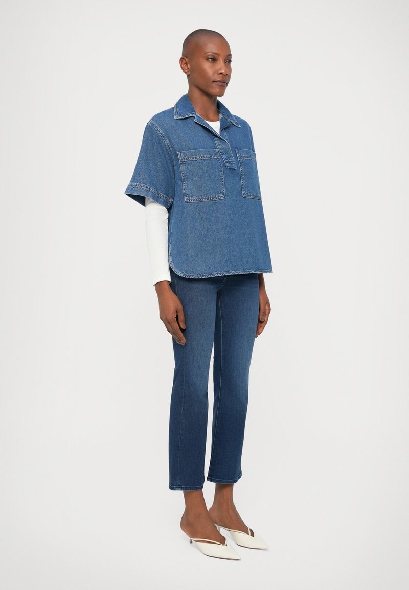 Jeanshemd in Blau, mit Kragen und kurzen Ärmeln, ausgestattet mit zwei Fronttaschen; kombiniert mit engen blauen Jeans und weißen Absatzmules.
