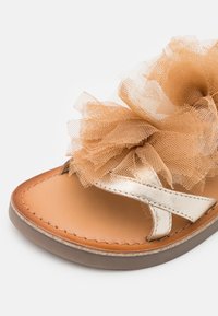 Brązowa skórzana sandał z metalowym paskiem i dużą, warstwową beżową aplikacją z tiulu na górze na białym tle.