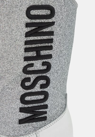 Botte en tissu argenté étincelant avec une broderie noire « MOSCHINO » audacieuse sur le côté et une semelle blanche lisse. Présente une vue rapprochée.