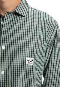 Grün-weißer kariertes Langarmhemd mit einer Brusttasche, die ein DC Shoes Logo-Patch trägt.