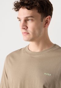 Kortærmet brun t-shirt lavet af bomuld, med rund halsudskæring og et lille grønt "HUGO" logo på venstre bryst. Glat tekstur.
