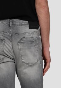 Jean en denim gris avec un effet délavé, deux poches arrière, une étiquette de logo noire sur la taille et des détails de couture visibles.