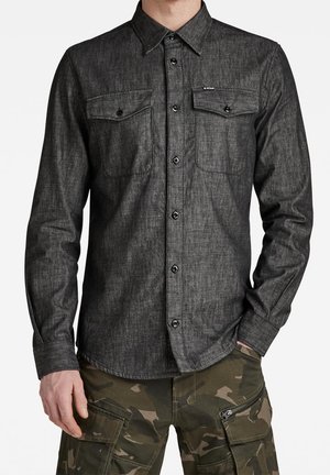Hombre con una camisa gris oscuro abotonada con dos bolsillos en el pecho y pantalones cargo de camuflaje con cremalleras y bolsillos con solapa.
