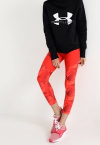 Leggings rouges à motifs en matériau extensible, associés à un hoodie noir avec un logo blanc et des chaussures de sport roses.