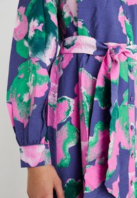 Blå klänning med stora rosa och gröna abstrakta blommönster. Har långa ärmar, midjeband och knäppning med knappar. Texturerat tyg.