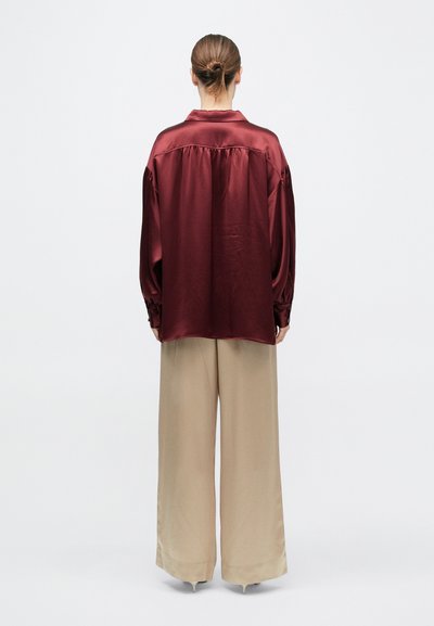 Person med brunt hår i en knold iført en løs bordeaux satinbluse og brede beige bukser, stående med ryggen til på en hvid baggrund.