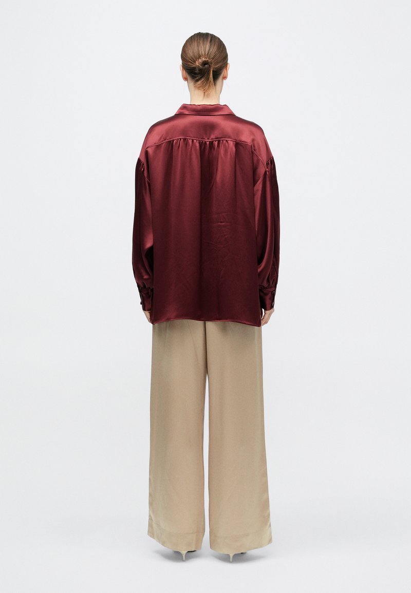 Person with brown hair in a bun wearing a loose burgundy satin shirt and wide beige trousers, standing facing away on a white background.