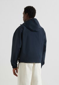 Veste imperméable bleu marine avec capuche, poignets élastiques et coupe décontractée. Caractéristiques d'une texture lisse et d'un design minimaliste.