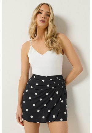 Vrouw met lang blond haar, gekleed in een witte tanktop en een zwarte wikkelrok met witte stippen, staand tegen een eenvoudige muur.