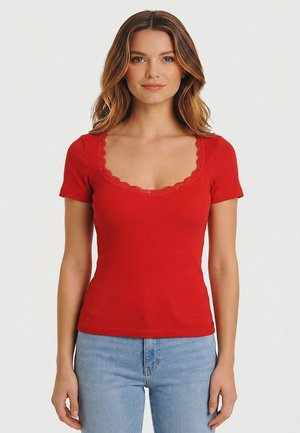 Donna che indossa una maglia rossa a maniche corte a costine con scollo in pizzo smerlato e jeans azzurri, in piedi contro uno sfondo bianco semplice.