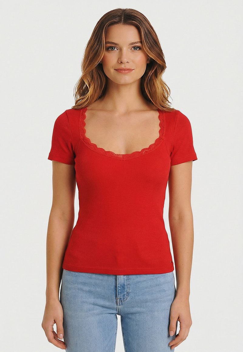 Donna che indossa una maglia rossa a maniche corte a costine con scollo in pizzo smerlato e jeans azzurri, in piedi contro uno sfondo bianco semplice.