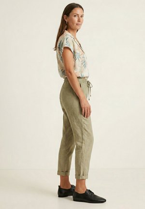 Kobieta ubrana w bluzkę z krótkim rękawem w tropikalny wzór, oliwkowe spodnie ze sznurkiem i czarne buty sznurowane, stoi bokiem na jednolitym tle.