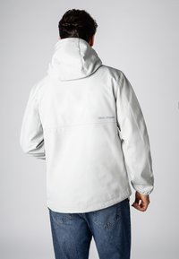 Helle graue Kapuzenjacke mit glatter Oberfläche, versehen mit einem Rückenlogo und elastischen Bündchen, kombiniert mit blauen Jeans.